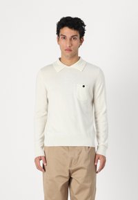 Pulôver de malha de cor creme com colarinho tipo polo, mangas longas e um bolso no peito com um botão preto. Tecido texturizado, design ajustado.