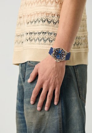 Marineblauwe rubberen horloge met een ronde zilveren kast, blauwe wijzerplaat en gouden accenten; gedragen om een hand tegen een gestructureerde beige gebreide top.
