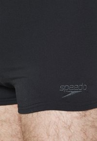 Sorte svømmebukser lavet af glat stof, med en tætsiddende design og et diskret gråt "Speedo"-logo på den nederste del.