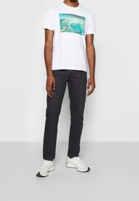 En person står självsäkert, iklädd en vit Levi's t-shirt med en färgglad grafik, mörka byxor och trendiga sneakers mot en enkel bakgrund.