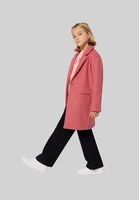 Manteau en mélange de laine rose avec des revers en notch, fermeture à un bouton, poches latérales, associé à un haut rose clair et un pantalon noir à jambes larges.