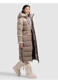 Palton lung bej din puf, cu glugă căptușită cu blană, fermoar frontal și buzunare laterale. Asortat cu cizme albe izolate. Textură netedă, matlasată.