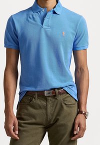 Man in een lichtblauwe polo met een klein oranje logo, olijfgroene broek, een bruine riem en een bruine leren horloge.