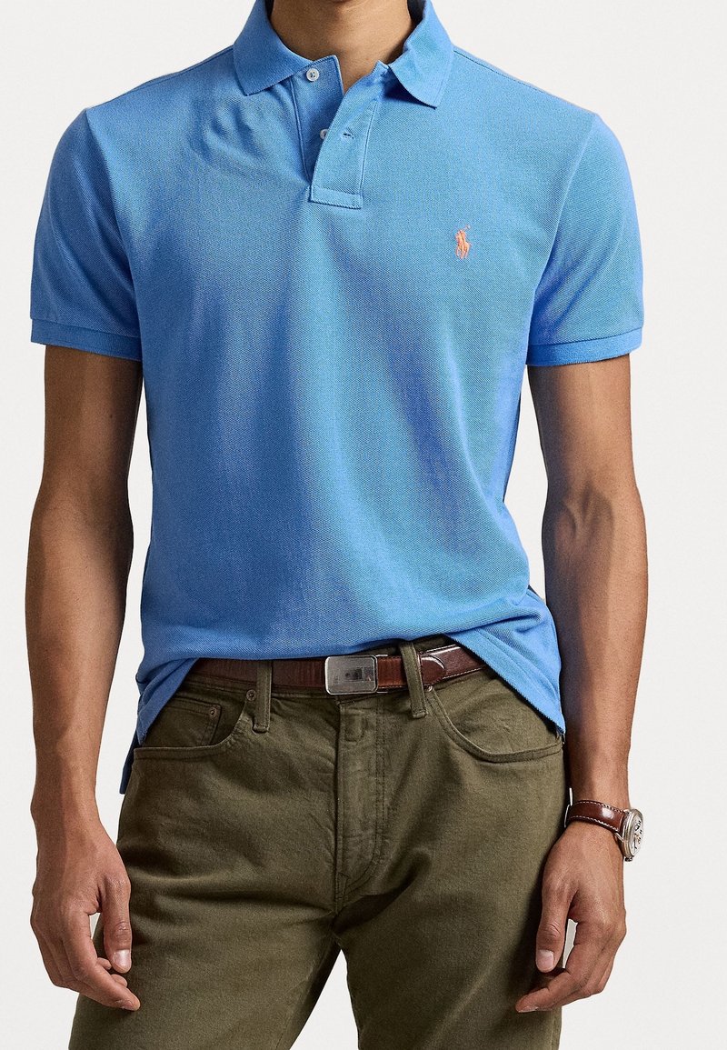 Man in een lichtblauwe polo met een klein oranje logo, olijfgroene broek, een bruine riem en een bruine leren horloge.