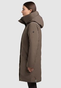 Bruine lange parka met een hoge kraag, voorzien van een ritssluiting en drukknoopsluiting, zijzakken en een zwart logo-accent op de mouw.