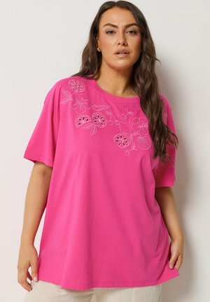 Femme aux longs cheveux bruns portant un t-shirt ample rose vif avec un motif linéaire blanc de fleurs et de fruits sur la poitrine, associé à un pantalon clair.