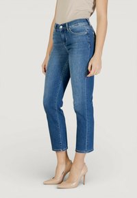 Femeie purtând blugi din denim albastru cu talie medie și pantofi cu toc înalt bej, stând pe un fundal simplu, deschis la culoare.