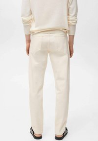Pantaloni color crema con vestibilità slim, realizzati in denim. Presentano tasche posteriori standard e dettagli cuciti. Indossati con un maglione leggero.