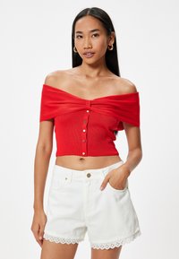 Rød off-shoulder crop top med ribbet tekstur og frontlukning med knapper, parret med hvide shorts med hulmønster i kanten og lommer.