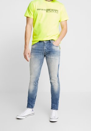 Camiseta amarilla neón con texto negro, combinada con jeans de denim azul con un lavado deslavado y detalles desgastados, usados con zapatillas blancas.
