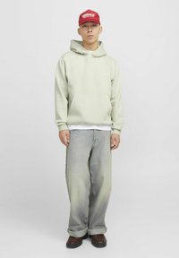 Hellgrüner Hoodie mit Kängurutasche, über einem weißen Shirt getragen; kombiniert mit lockeren, ausgebleichten Jeans und braunen Schuhen; rotes Cap.