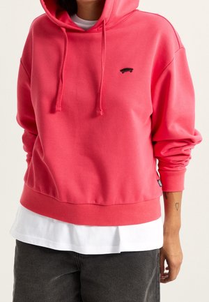Personne portant un sweat à capuche rose avec un petit logo skateboard noir, une chemise blanche superposée en dessous et un pantalon noir.