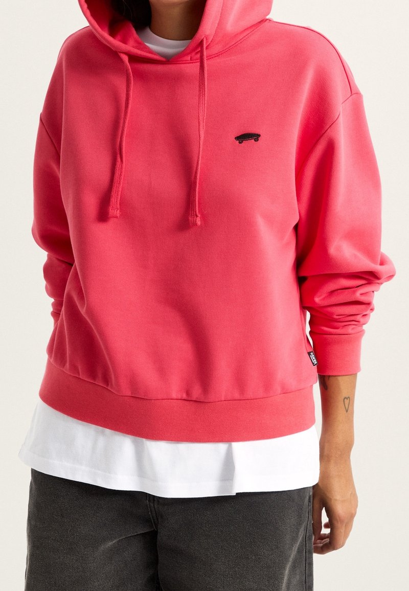Personne portant un sweat à capuche rose avec un petit logo skateboard noir, une chemise blanche superposée en dessous et un pantalon noir.