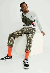 Homem sentado em um cubo branco, vestindo calças camufladas, sweatshirt cinza, bolsa de ombro verde, meias laranja da Vans e tênis pretos da Vans.