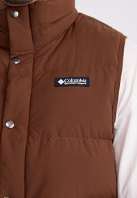 Smeđa izolirana prsluk s visokim ovratnikom, metalnim kopčama i crnim tekstilnim logom koji prikazuje "Columbia Sportswear Company."