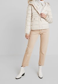 Giubbotto imbottito crema con cappuccio e rifinitura in pelliccia, polsini a coste, zip frontale e tasche laterali. Abbinato a pantaloni beige e stivali alla caviglia bianchi.