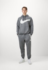 Felpa Nike grigia con un grande logo bianco, abbinata a pantaloni tuta grigi coordinati con un accento bianco. Le scarpe sono bianche, con una finitura testurizzata.