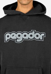 Μαύρη κουκούλα με μεγάλο γκρίζο λογότυπο "pegador" μπροστά. Διαθέτει τσέπη καγκουρό και κουκούλα με κορδόνι. Μαλακή υφή υφάσματος.