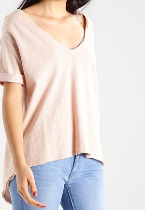 Frau trägt ein locker sitzendes, blassrosa V-Ausschnitt-T-Shirt mit hochgekrempelten Ärmeln und hellblauen Jeans, gezeigt von Hals bis Hüfte.