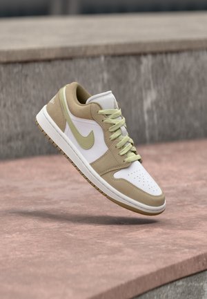 Beige and white Nike sneaker with light green laces and swoosh, shown mid-air over pink concrete steps.