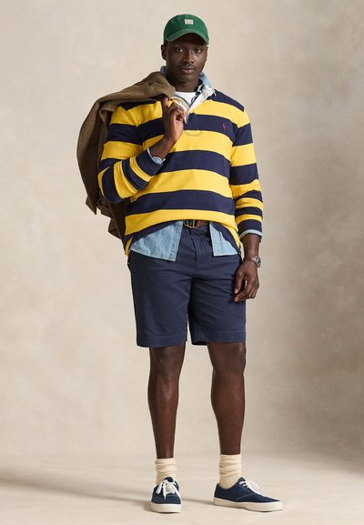 Man wearing green cap, yellow and navy striped long-sleeve shirt, denim shirt, navy shorts, cream socks, navy sneakers, holding brown jacket over shoulder.