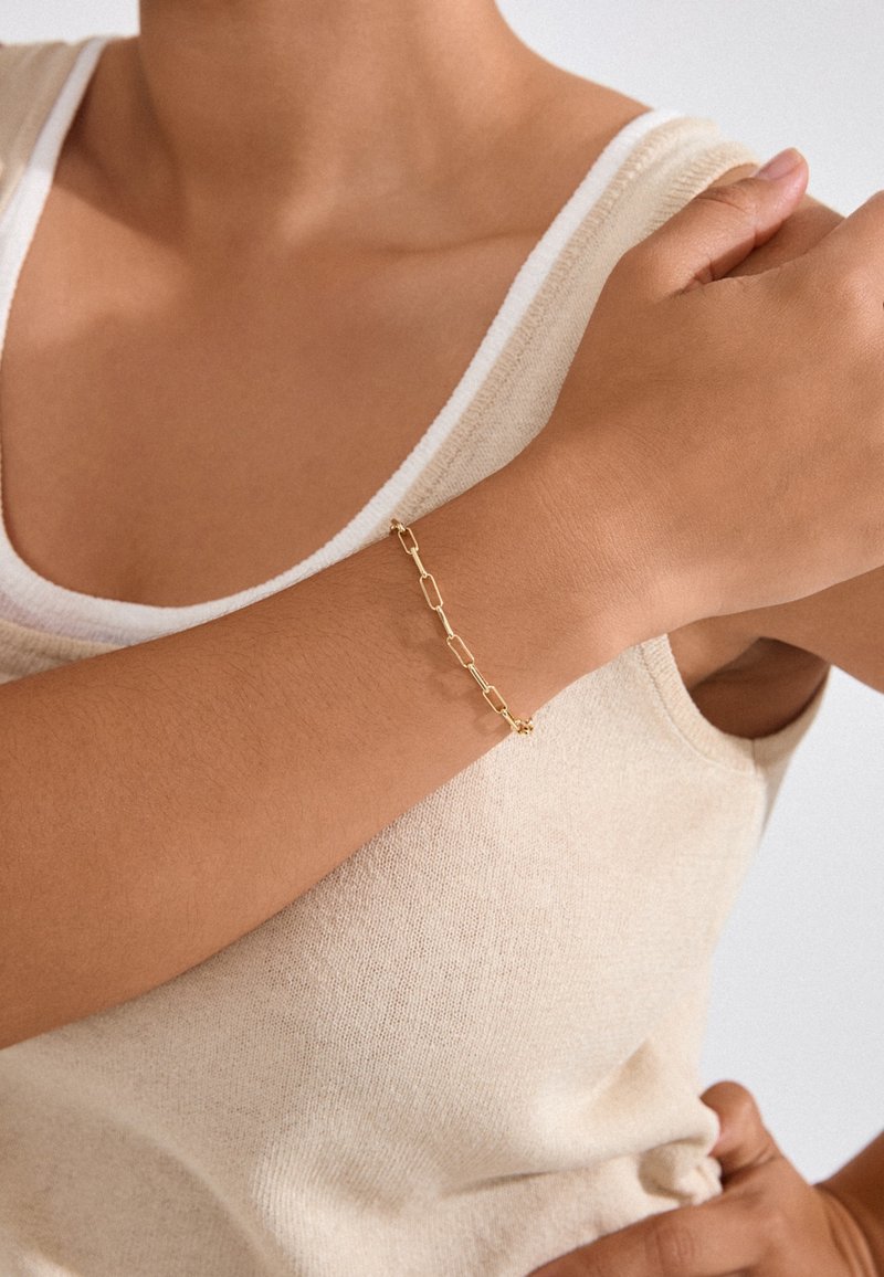 Kuldne kett käevõru, millel on ristkülikukujulised lingid ja väike tähedetail, kantud paljal randmel tekstuurse beeži sviitri vastu.