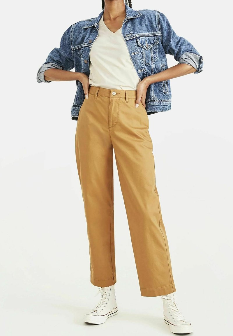Femme portant une veste en jean retroussée, un t-shirt blanc à col en V, un pantalon taille haute couleur beige, et des baskets blanches montantes, debout, les mains sur les hanches.