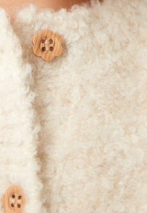 Gros plan sur un pull beige et duveteux avec deux boutons en forme de fleurs en bois attachés à l'avant.