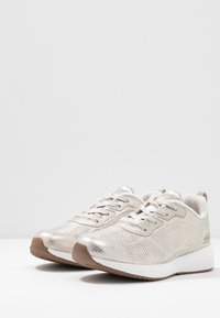 Zapatillas atléticas de color plata metálico con un acabado texturizado, diseño con cordones y una suela de goma blanca con un agarre estampado.