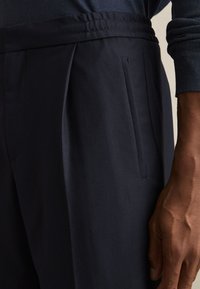 Pantalones a medida de color azul marino oscuro con bolsillos laterales, combinados con una camiseta de manga larga a juego en azul marino oscuro, mostrados de lado derecho en la cintura y la mano.