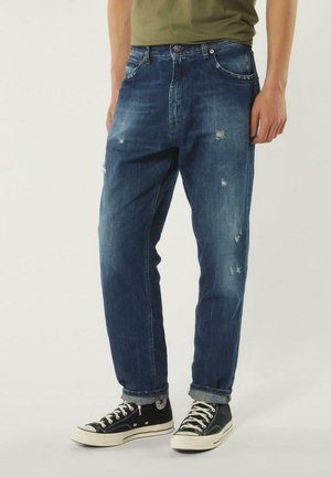 Jeans in denim blu scuro con toppe strappate, design a cinque tasche, vestibilità dritta e orli arrotolati. Abbinati a sneaker nere.