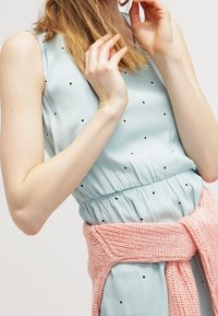 Vaaleansininen hihaton mekko mustilla pilkuilla, joustava vyötärö, yhdistettynä pinkkiin neulottuun vyötäröliinaan. Sileä kankaan rakenne.