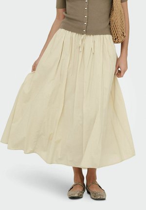 Femme portant un haut beige boutonné avec une jupe longue crème, tenant la jupe et portant un sac en paille tissé à l'épaule.