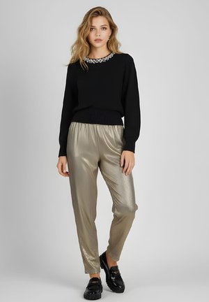 Mujer con un suéter negro con escote adornado con joyas, pantalones metálicos dorados y mocasines negros brillantes, de pie frente a un fondo gris.