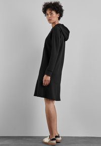 Robe noire à capuche en tissu doux, à manches longues, avec une coupe décontractée et un ourlet qui tombe au milieu de la cuisse, assortie à des baskets beiges.