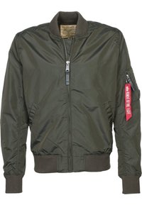 Veste bomber verte avec fermeture éclair à l'avant, poignets et bas côtelés, et étiquette rouge « Remove Before Flight » sur la fermeture éclair de la poche de la manche gauche.
