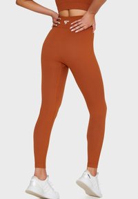 Prążkowane, przylegające legginsy w kolorze spalonej pomarańczy, z wysokim pasem i białym logo. W połączeniu z białymi butami sportowymi.