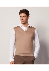 Beżowy sweter bez rękawów z dekoltem w kształcie V, noszony na białej koszuli z kołnierzykiem, tworzący warstwowy wygląd; gładka tekstura i dopasowany krój.