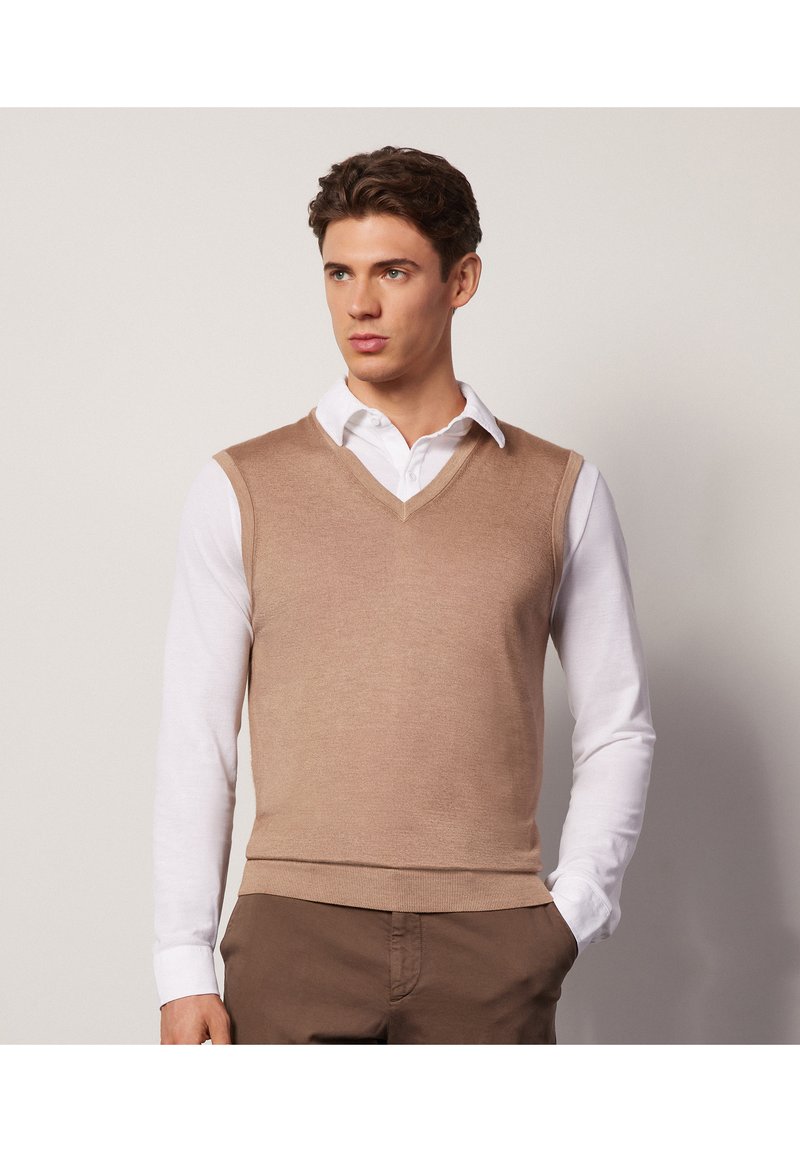 Beżowy sweter bez rękawów z dekoltem w kształcie V, noszony na białej koszuli z kołnierzykiem, tworzący warstwowy wygląd; gładka tekstura i dopasowany krój.