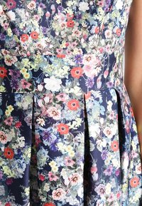 Blumendruckkleid mit navy Hintergrund, das bunte Blumen zeigt. Faltenwurf-Design mit weichem Stoff. 