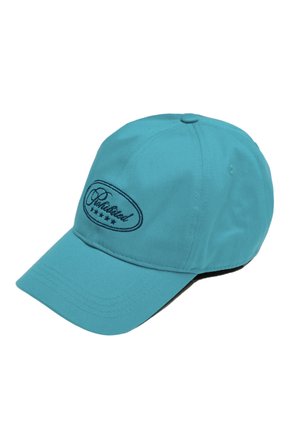 Une femme porte une casquette de baseball turquoise brodée, un haut sans manches côtelé couleur crème, des créoles dorées et un collier à pendentif, sur un fond uni.