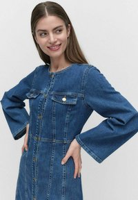 Kangashame, jossa on pyöreä kaula-aukko, nappisulku, kaksi rintataskua ja levenevät hihat. Kangas on keski sinisen pessun värinen, jossa on haaleita ompeleita.