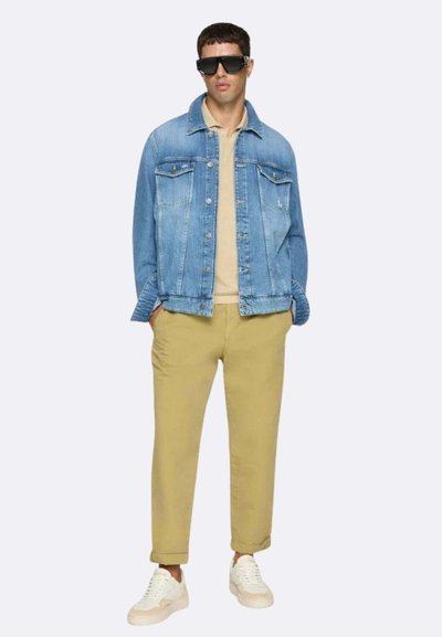 Giacca di jeans azzurro chiaro, polo beige, pantaloni kaki e sneakers bianche. Occhiali da sole e un fit rilassato con dettagli di cucitura visibili.