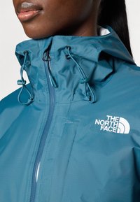 Prim-plan cu o persoană care poartă o geacă impermeabilă The North Face albastră, cu glugă și șnururi reglabile, fermoarul tras până la gât.