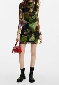 Robe moulante à manches longues avec motif abstrait multicolore, présentant des détails froncés. Associée à un petit sac à main rouge et des bottines noires.