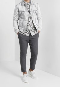 Personne portant une veste en denim clair, une chemise à motifs floraux avec des boutons, un pantalon gris foncé et des chaussures blanches à lacets, les mains dans les poches.