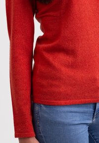 Röd stickad tröja med mjuk textur, långa ärmar och rak nederkant, ihop med blåjeans. Inga iögonfallande mönster eller detaljer.