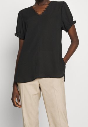 Dark-skinned person wearing a black short-sleeve blouse with lace trim and beige pinstriped pants, posing with one hand in pocket.