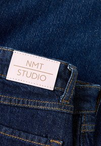 Tummansininen denimkangas, jossa on kultaiset tikit. Valkoisessa lapussa on kultaisella tekstillä "NMT STUDIO", ja se on kiinnitetty vyötärölle.