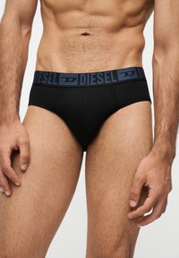 Buste masculin portant un slip Diesel noir avec ceinture de marque, abdomen et cuisses visibles, mains détendues sur les côtés.